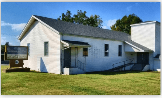 Exterior of a white, single-story church building with a sign in front. Green lawn surrounds the structure, indicating a serene community setting.