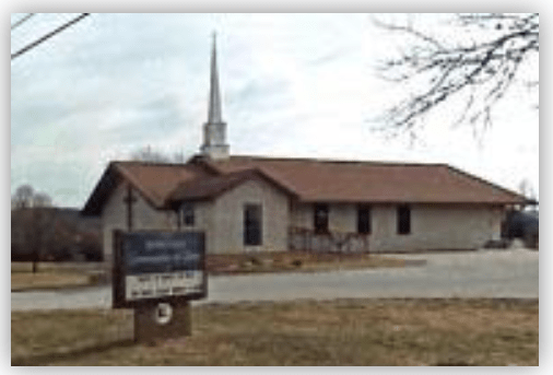 A modest church building with a steeple, featuring a sign in front. The image highlights its welcoming presence in the community.
