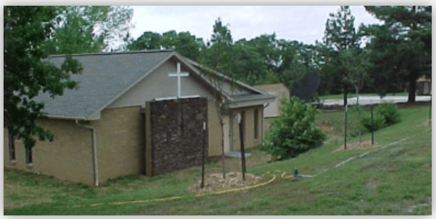 Small church exterior featuring a prominent cross, surrounded by greenery and a freshly landscaped area, suggesting community engagement or renovations.