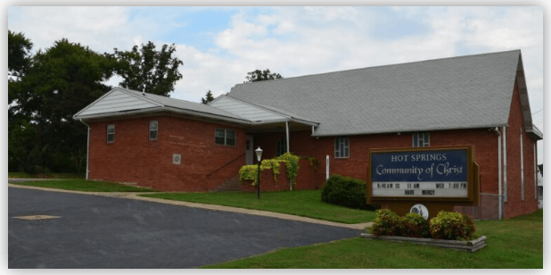 Community of Christ church building in Hot Springs, featuring a brick exterior and a sign listing service times. Lush green landscaping surrounds the entrance.