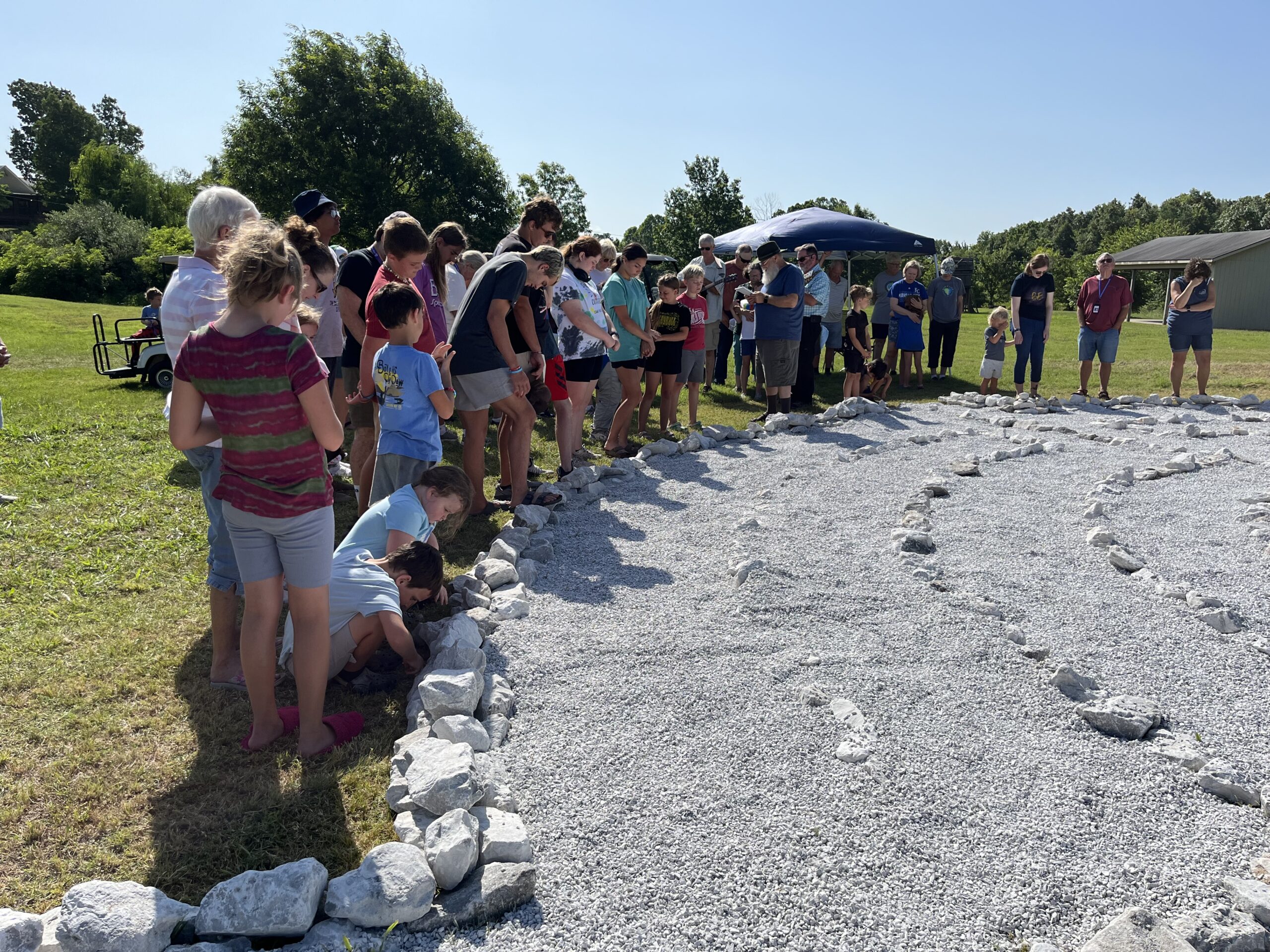 A diverse group of people, including children, gather around a newly created rock pathway in a grassy area, enjoying a community event under bright skies.