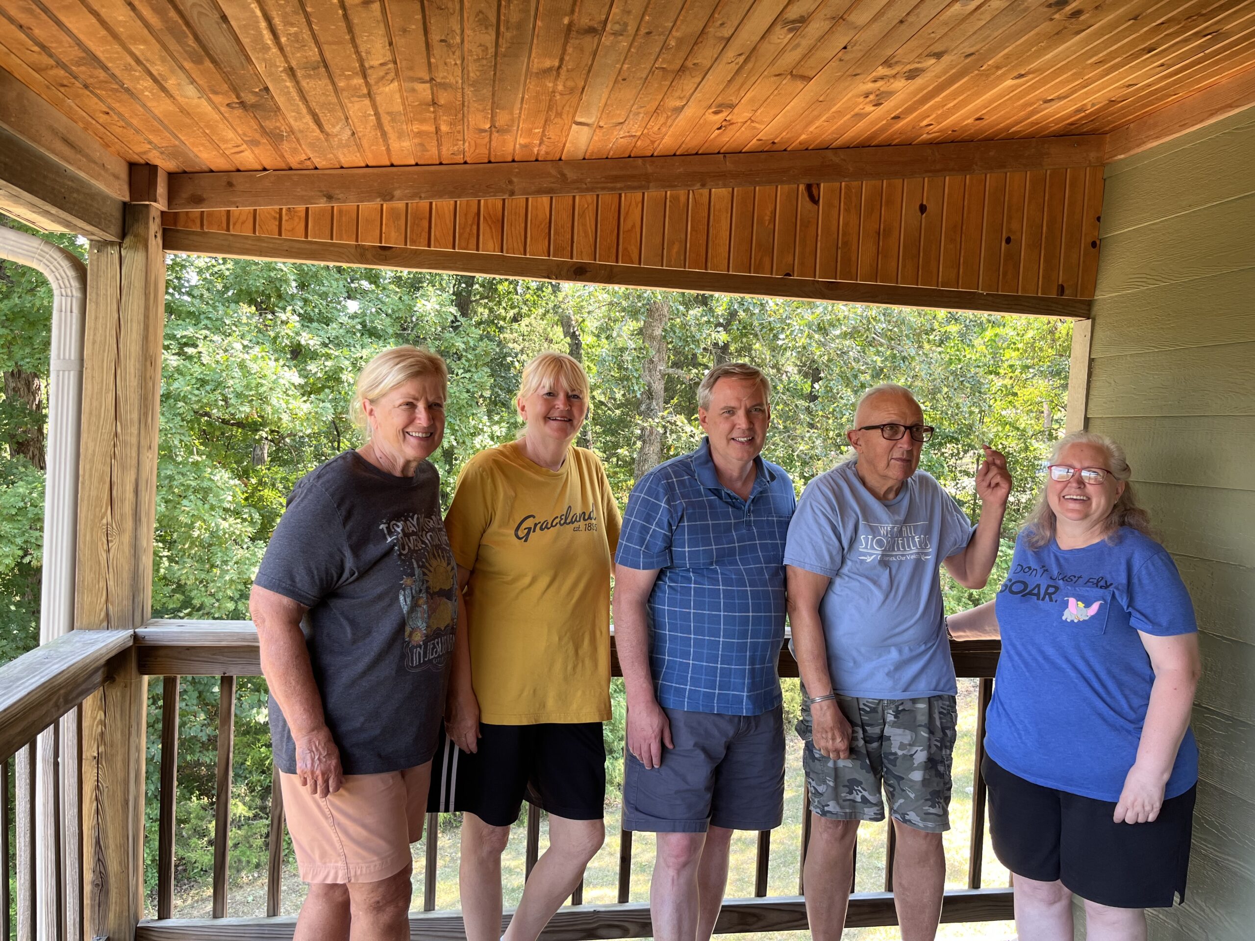 Five friends stand together on a wooden porch surrounded by greenery, smiling and enjoying a sunny day. The image captures a moment of camaraderie.