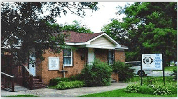 Brick building with white trim, housing the Kids Outdoor Bible Club, surrounded by greenery and a sign displaying its name.