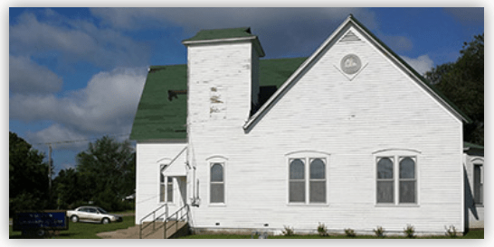 Historic white church with a green roof, featuring large windows and a front staircase, surrounded by trees and a parked car.