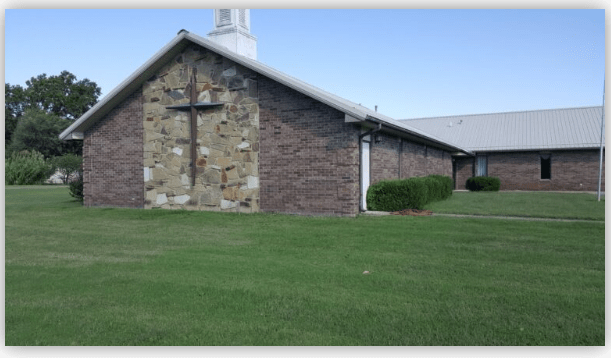 Brick church exterior featuring a stone wall with a prominent cross. Lush green lawn surrounds the building, emphasizing its inviting atmosphere.