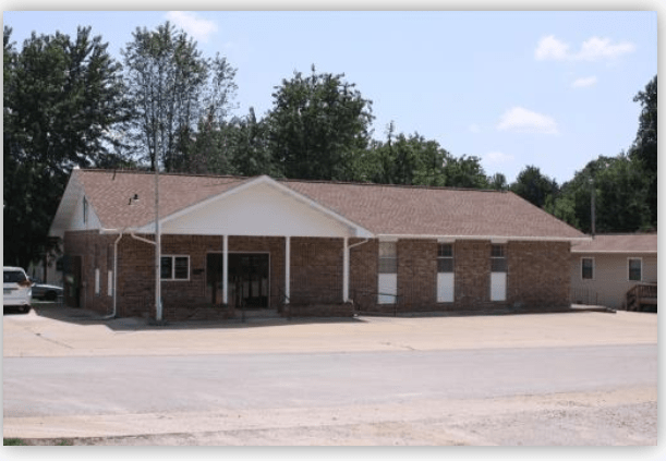 Brick building with a sloped roof and front porch, surrounded by trees. Represents a community space or facility in a rural area.
