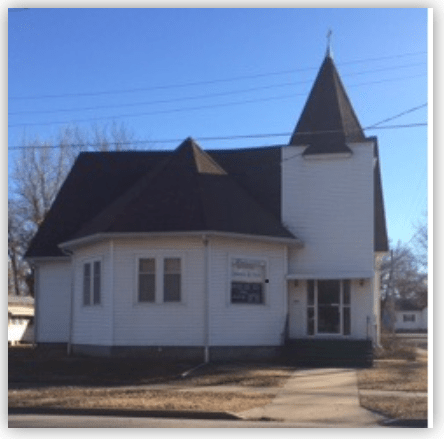 An eclectic church building with a pointed steeple, featuring a white exterior and large windows, set against a clear blue sky.