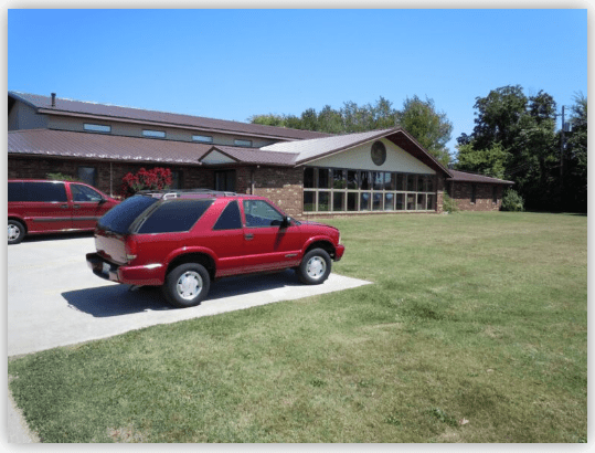 Red SUV parked in front of a large brick building with multiple windows and a well-kept lawn, indicating a welcoming facility or residence.