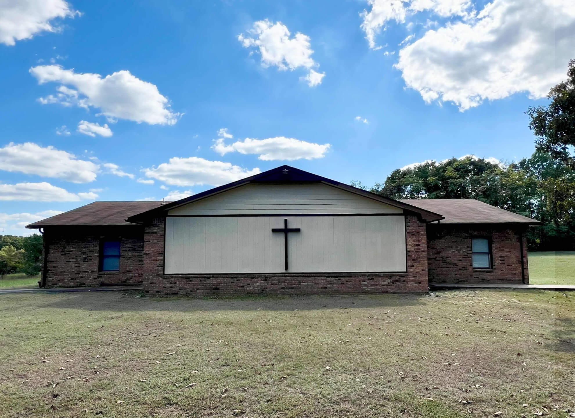 Brick church building with a large central cross on the facade, set against a blue sky with clouds. Surrounding area features grassy fields.
